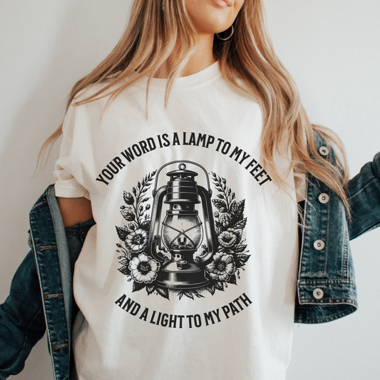 women in a crewneck short sleeve christian bible verse tee that says your word is a lamp to my feet and a light to my path with a vintage oil lantern and flowers