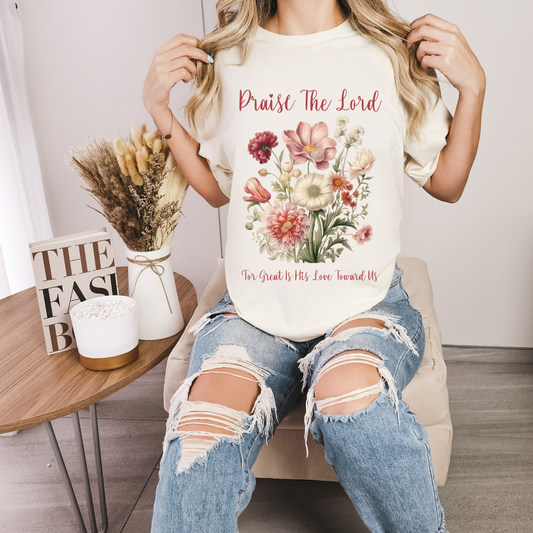 woman sitting in a chair wearing a womens christian t shirt that has a floral design with a bible verse that says praise the lord for great is his love towards us